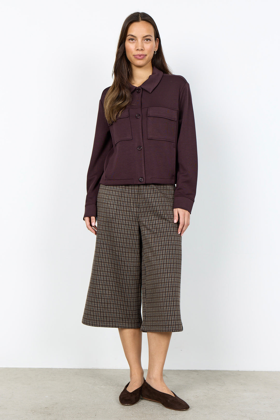 A woman stands against a plain white studio wall wearing a dark plum button-front jacket with flap chest pockets, brown and beige plaid cropped wide-leg pants, and dark brown slip-on shoes.