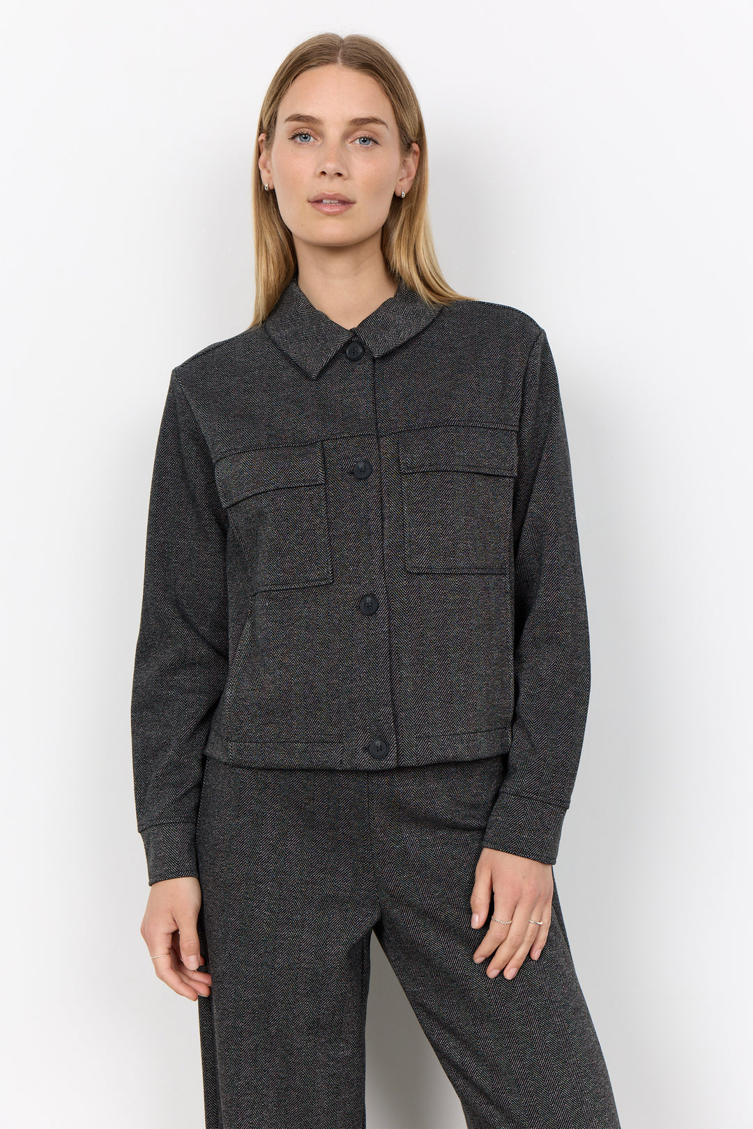 A woman modeling a dark gray textured jacket with a pointed collar and front button closure, paired with matching dark gray trousers, against a plain light gray studio background.