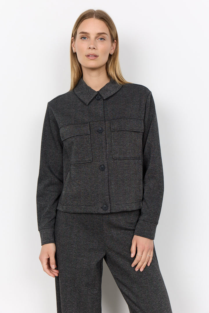 A woman modeling a dark gray textured jacket with a pointed collar and front button closure, paired with matching dark gray trousers, against a plain light gray studio background.