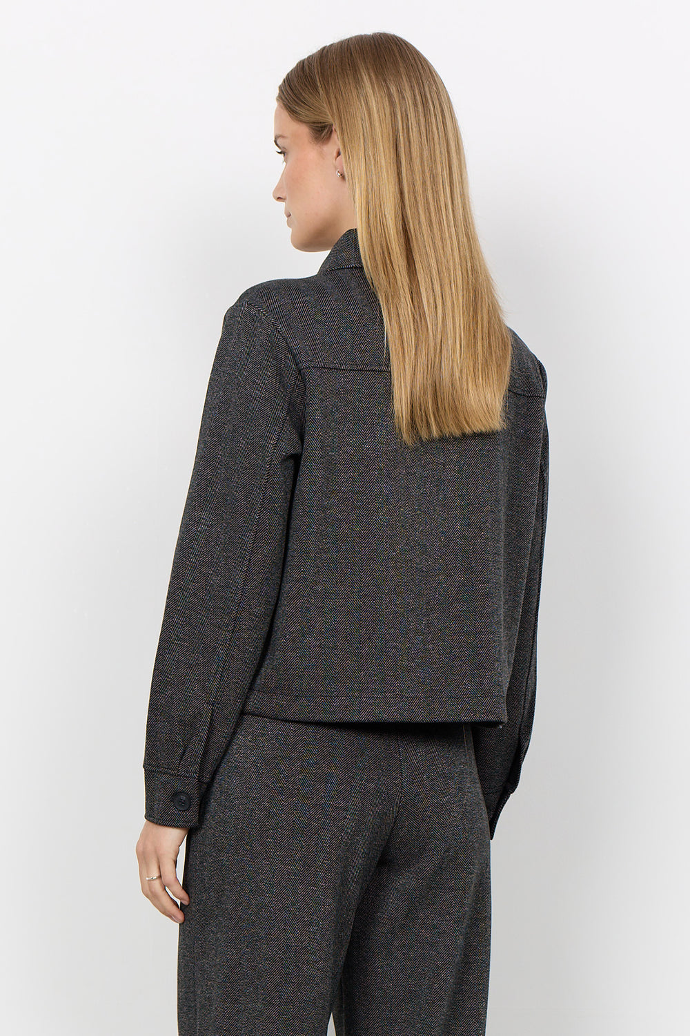 A woman with long blonde hair is shown from the back wearing a dark gray tweed cropped jacket and matching trousers against a plain white studio background.