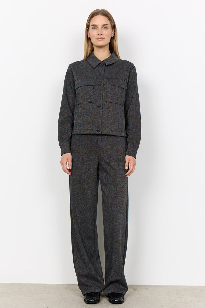 A woman stands against a plain white wall wearing a dark gray textured two-piece set consisting of a cropped button-front jacket with a pointed collar and matching wide-leg trousers.