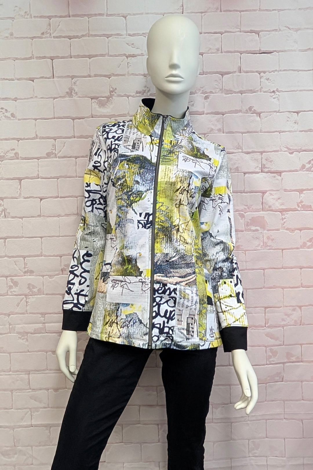 A long-sleeve zip-up jacket with a complex abstract graffiti and collage print in yellow, black, white, and gray, displayed on a female mannequin against a light pink brick wall.