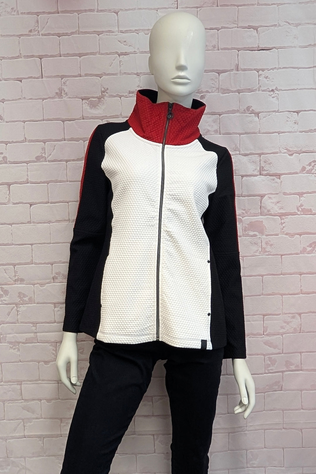 A mannequin wearing a  long-sleeve athletic zip-up jacket with a black and white color-blocked design, featuring red stripe accents on the sleeves, shown against a white brick background.