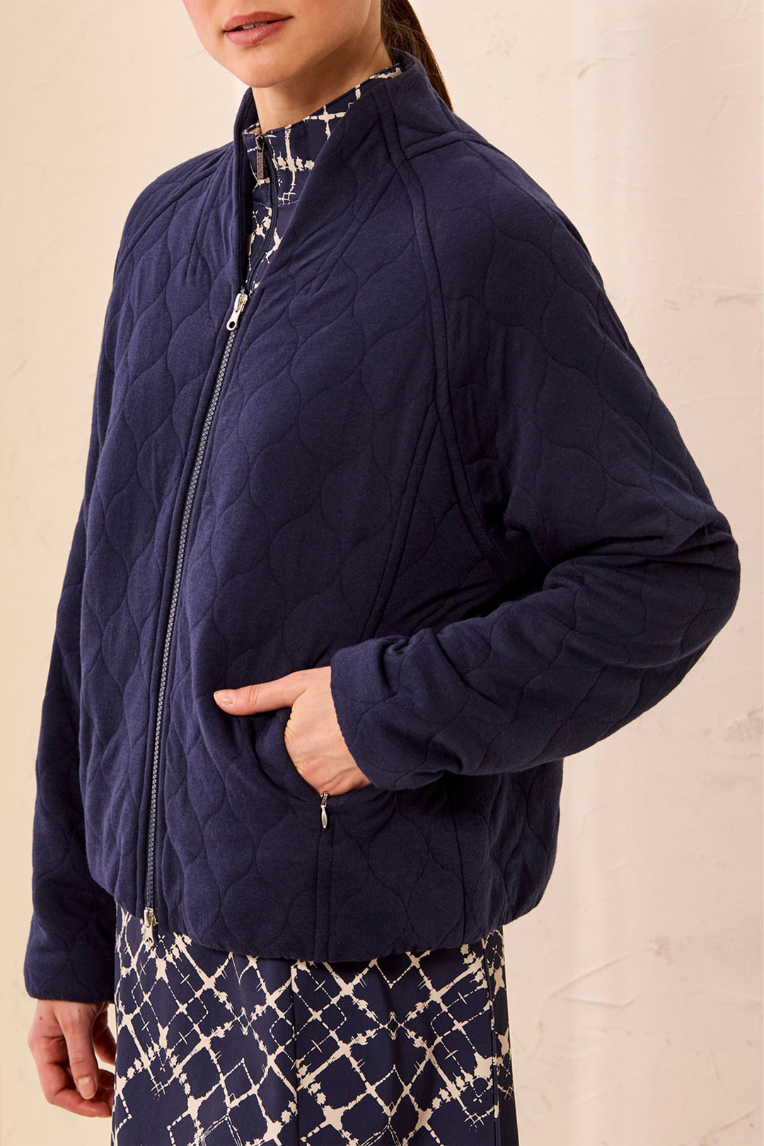 A woman wearing a navy blue quilted zip-up jacket over a patterned dress, shown from the neck down against a light beige textured wall.