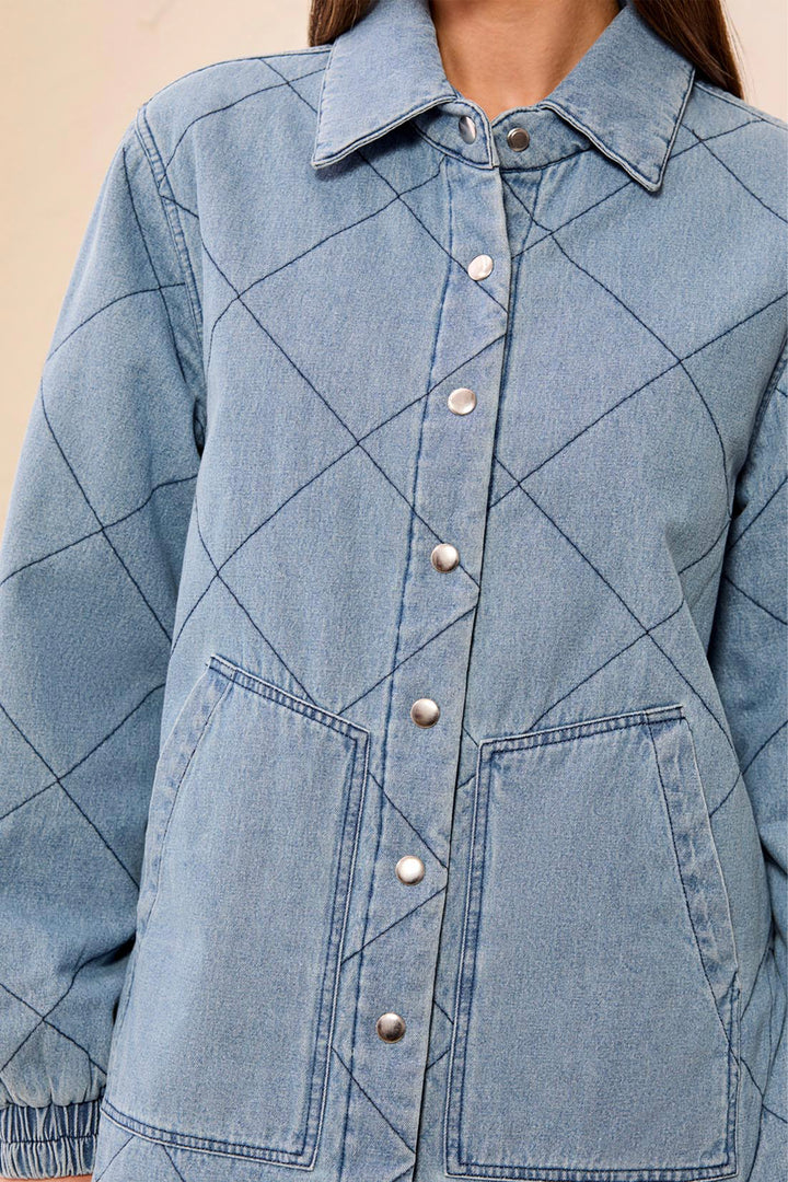 A close-up of a person wearing a light blue denim jacket with a diamond quilted pattern, featuring a pointed collar, silver snap buttons, and two front patch pockets.