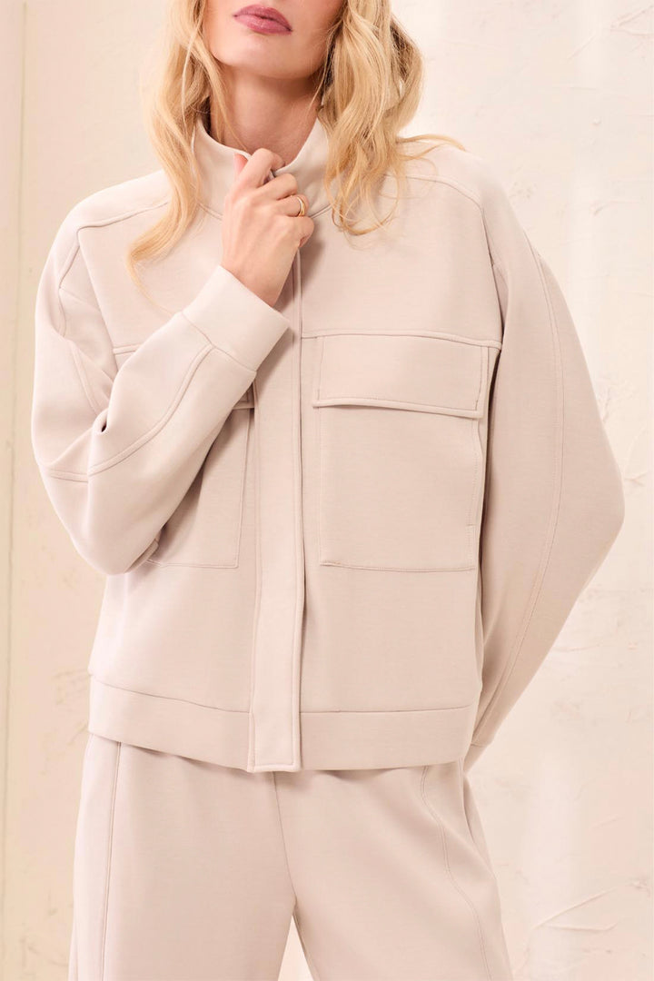 A woman wearing a light beige cropped jacket with a mock neck and flap pockets, paired with matching beige trousers, set against a soft beige background.
