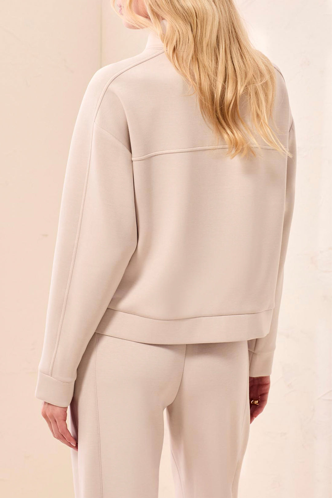 A woman shown from the back wearing a light beige cropped sweatshirt and matching sweatpants against a soft beige studio background.