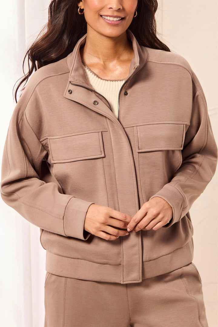 A woman wearing a taupe-brown utility jacket with a stand-up collar, snap-button placket, and two large flap chest pockets, paired with matching pants and a cream ribbed top.