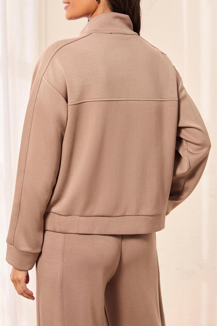 A person shown from the back wearing a taupe-beige cropped jacket and matching pants against a soft cream curtain background.