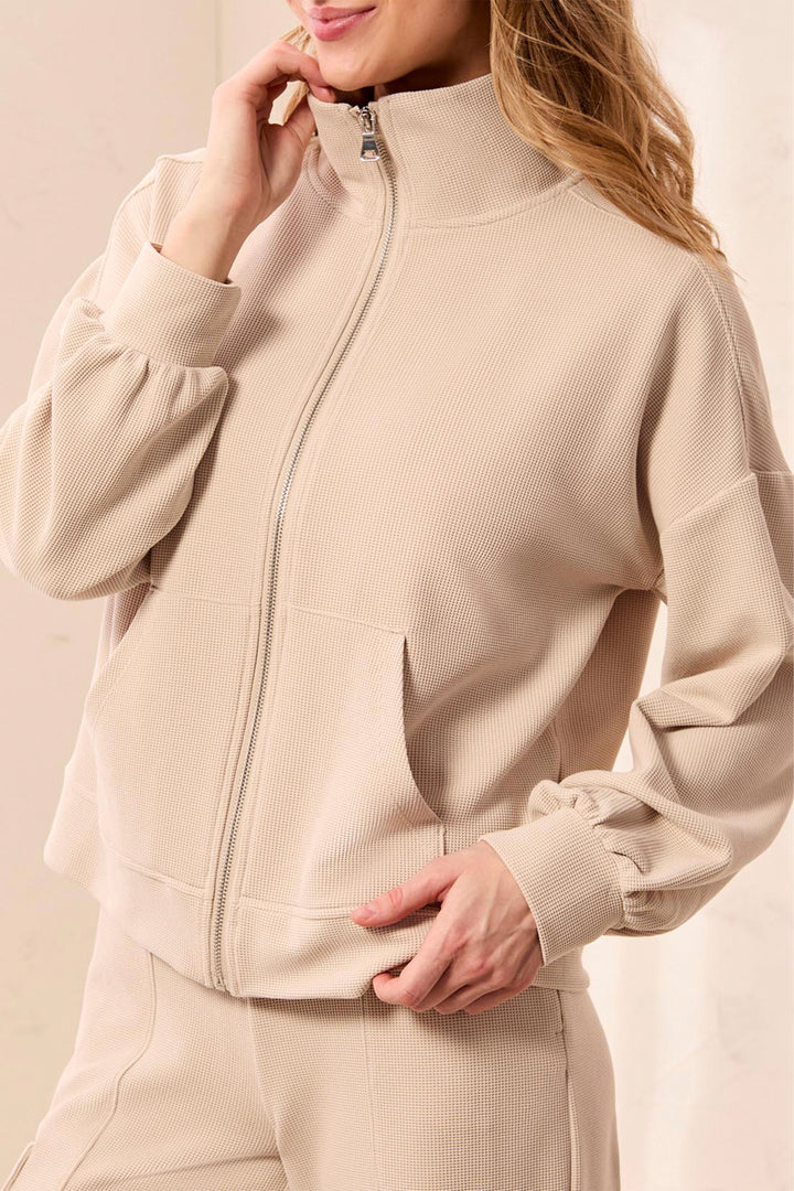 A woman wearing a beige ribbed knit zip-up jacket with long balloon sleeves and matching pants, shown in a close-up studio shot.