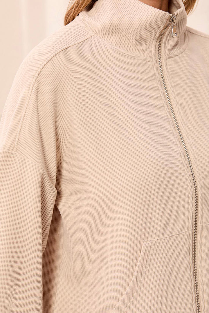 Close-up of a person wearing a light beige waffle-knit zip-up jacket with a high mock neck collar and a front zipper.