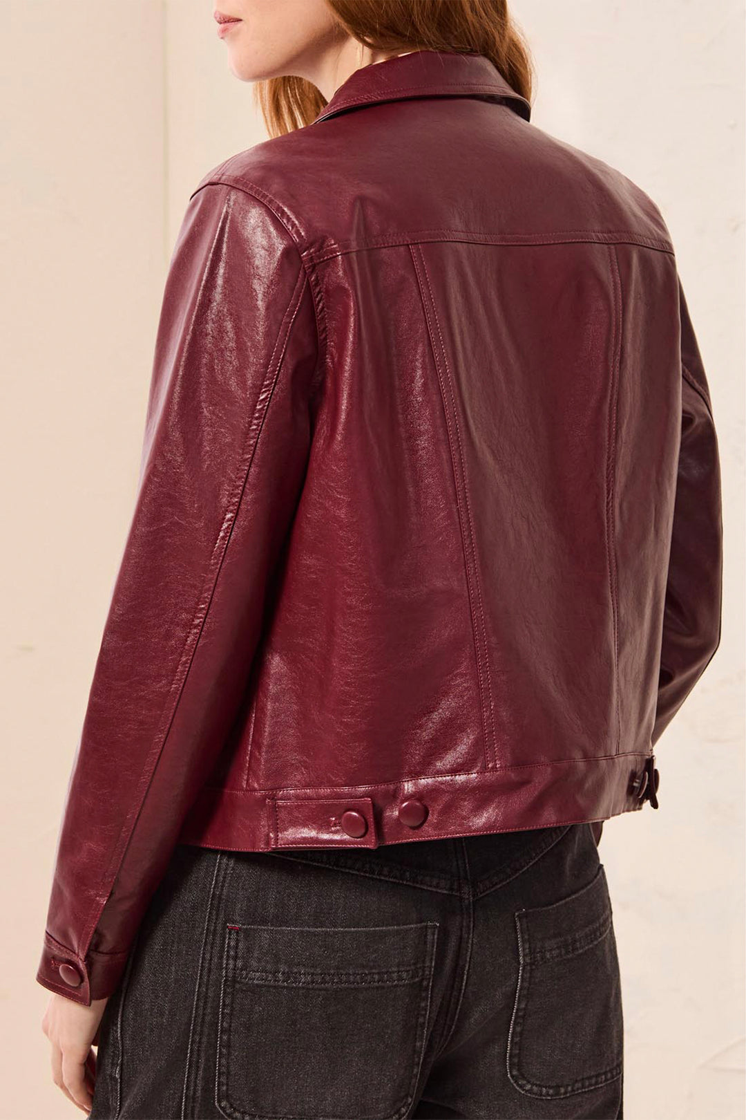 A person wearing a cropped, glossy burgundy leather jacket, shown from the back against a plain light beige background.