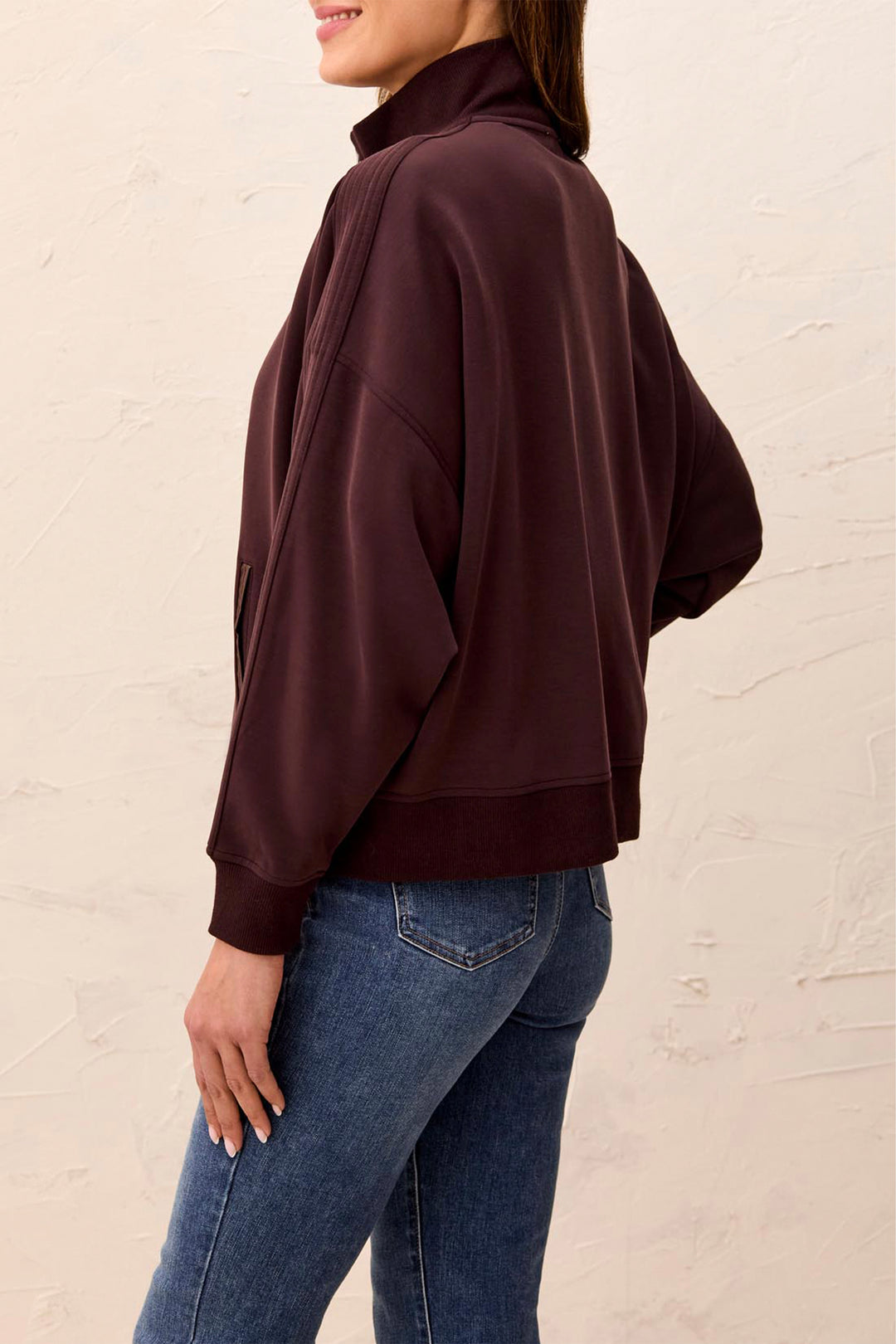A woman wearing a dark plum-brown cropped quarter-zip sweatshirt and blue jeans, shown from a side-back angle against a textured off-white wall.