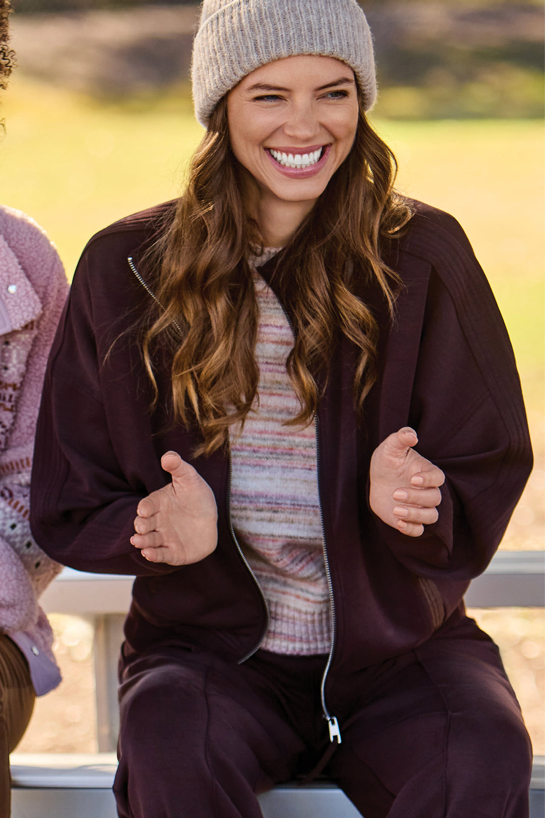 A woman outdoors wearing a matching dark plum zip-up jacket and pants set with a striped knit top and a beige beanie.