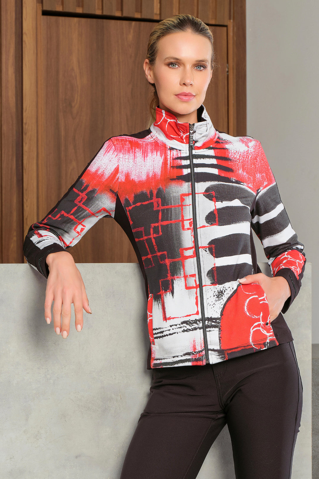 A woman modeling a long-sleeve athletic zip-up jacket with a bold abstract pattern in red, black, and white, paired with black leggings, posed in front of a wooden door and concrete wall.