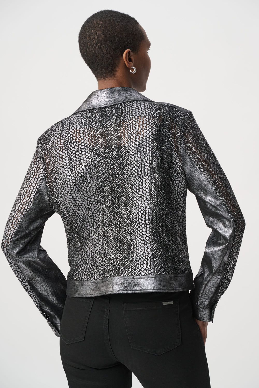 A woman shown from the back wearing a dark silver moto style jacket, and black pants in front of a white background. 