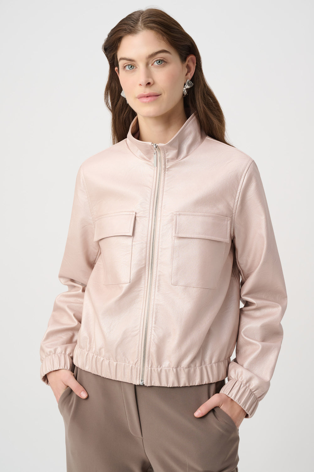 A woman wearing a light blush pink zip-up jacket with a stand collar and two front flap pockets, paired with taupe trousers, against a light gray studio background.