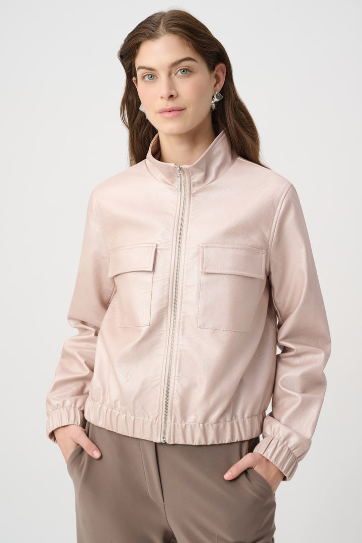 A woman wearing a light blush pink zip-up jacket with a stand collar and two front flap pockets, paired with taupe trousers, against a light gray studio background.