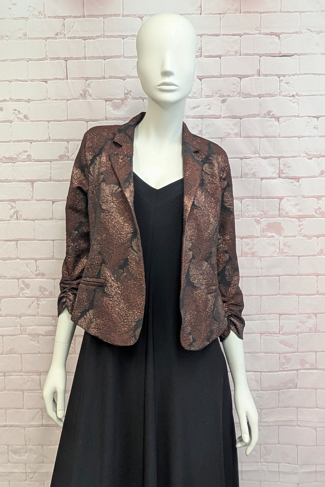 A mannequin standing in front of a light brick wall wearing a metallic patterned blazer. 
