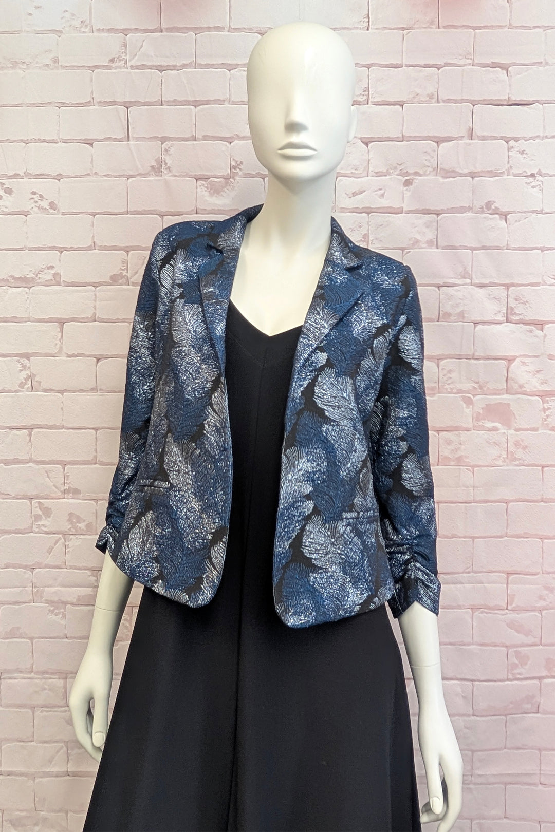 A mannequin standing in front of a light brick wall wearing a metallic patterned blazer. 