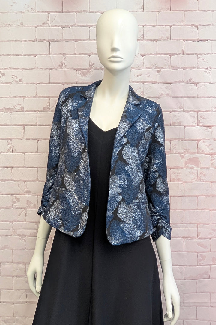 A mannequin standing in front of a light brick wall wearing a metallic patterned blazer. 