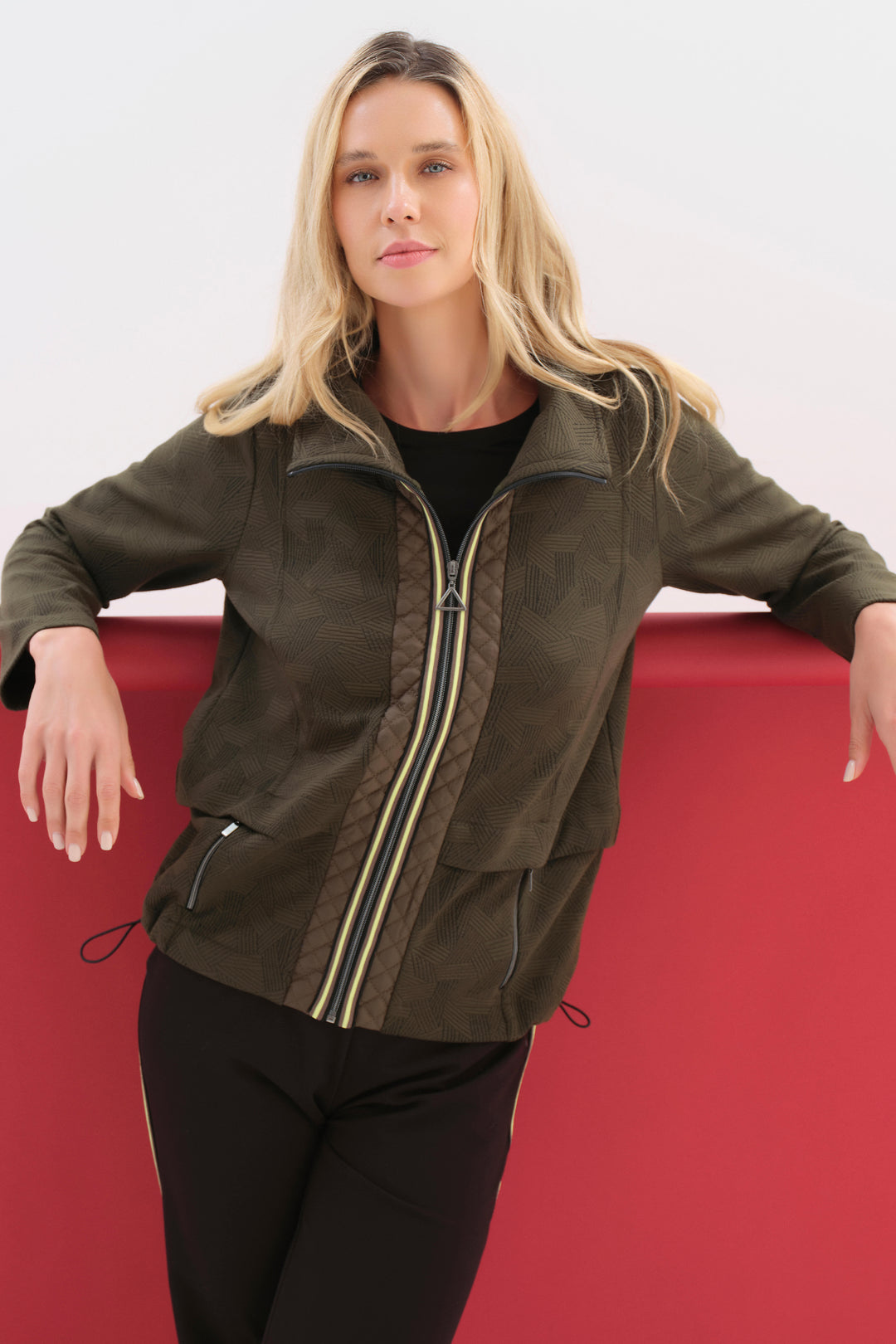 A woman wearing an olive green zip-up jacket with quilted brown patterned panels and gold-toned zipper accents, posed against a red and white studio background.
