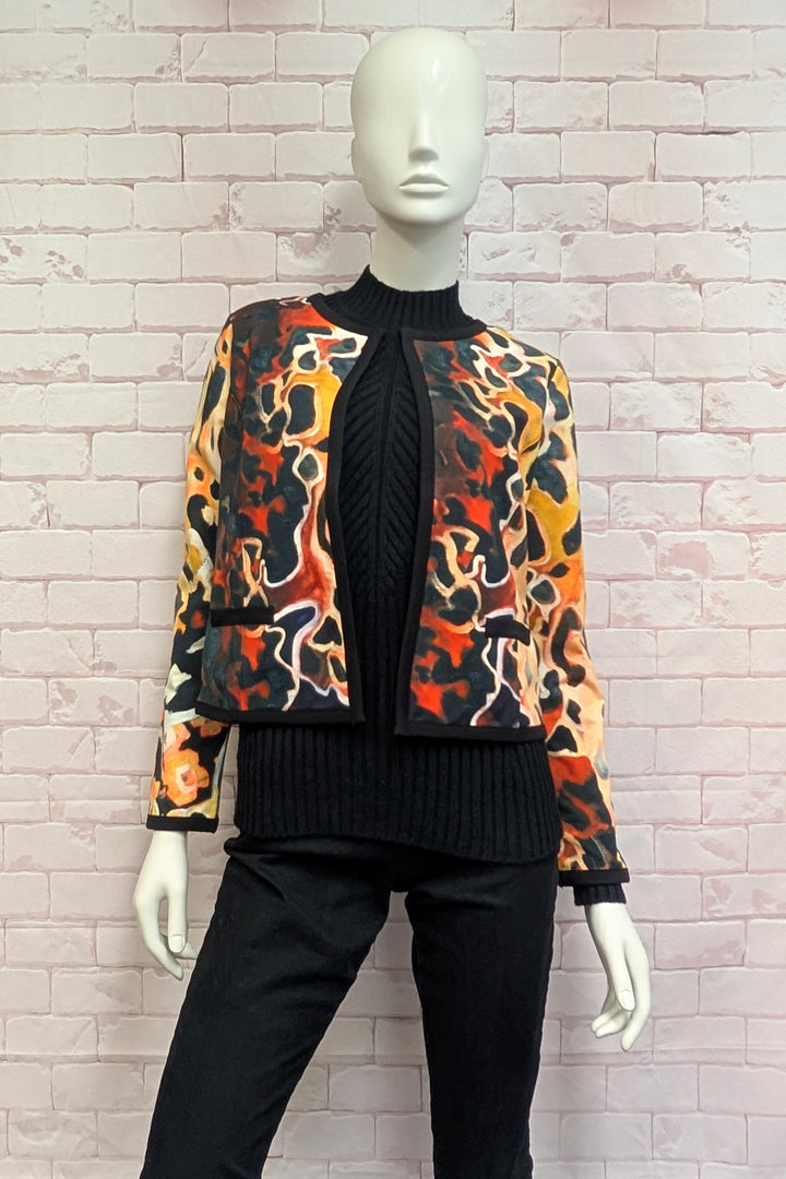 A mannequin wearing a cropped long-sleeve jacket featuring a bold abstract camouflage pattern in orange, black, beige, and yellow, with a black center front panel and trim.