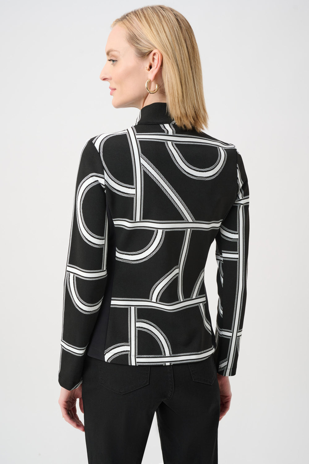 A woman shown from the back standing in front of a light grey background wearing a black and white geometric print jacket and black pants. 