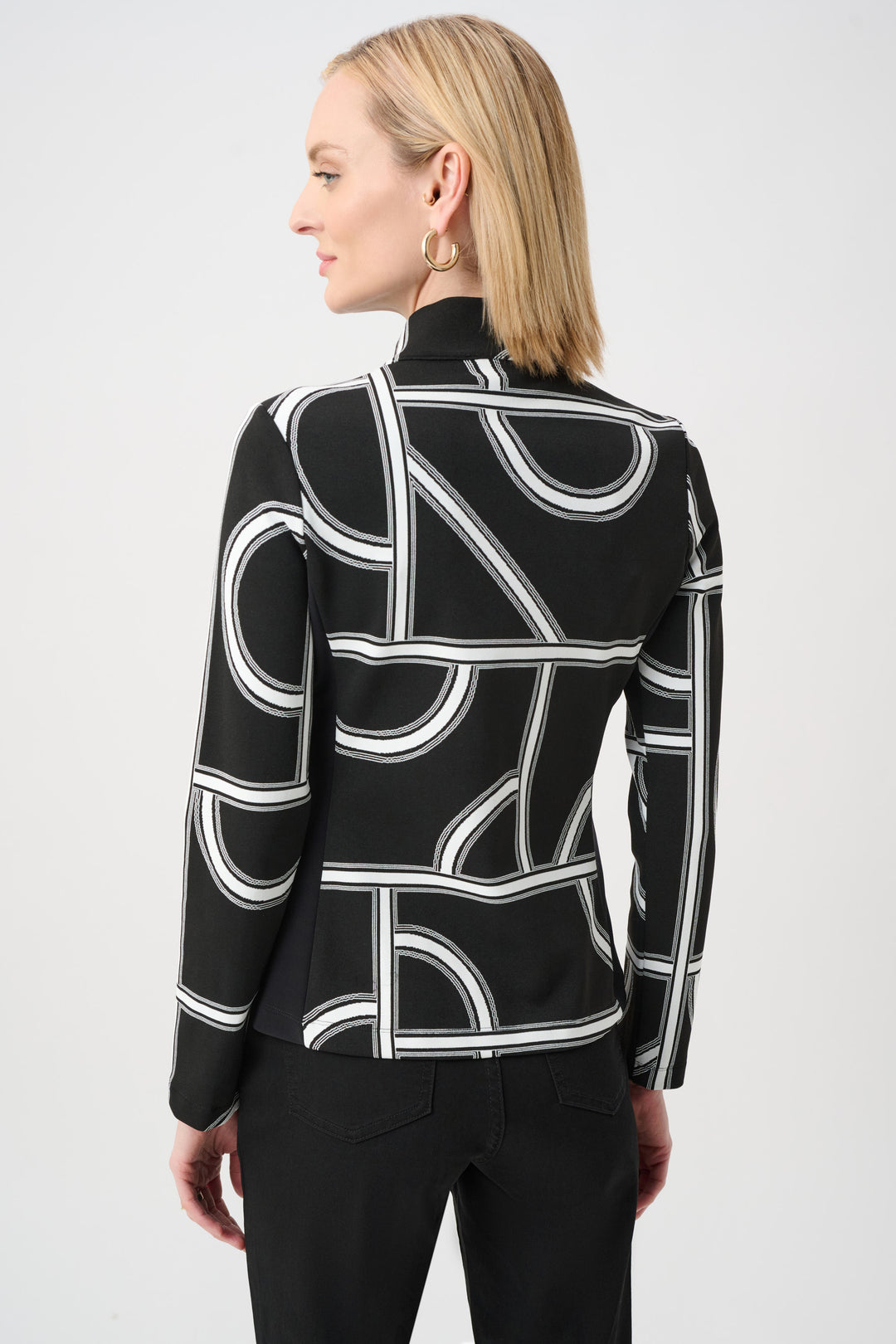 A woman shown from the back standing in front of a light grey background wearing a black and white geometric print jacket and black pants. 