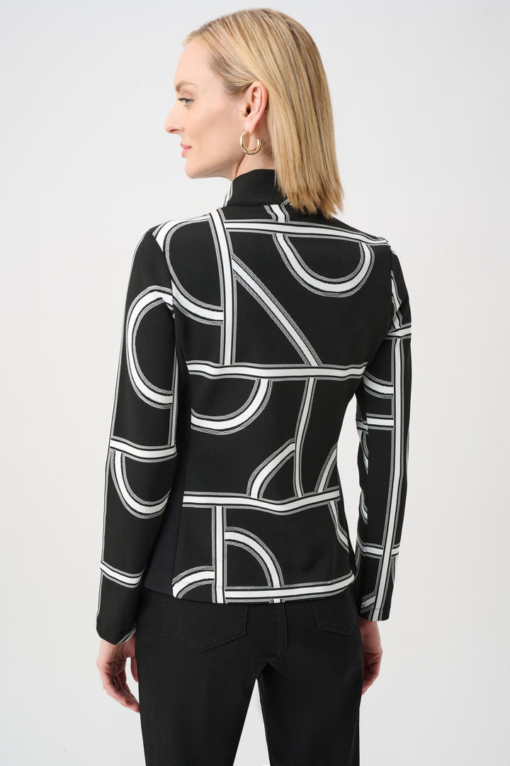 A woman shown from the back standing in front of a light grey background wearing a black and white geometric print jacket and black pants. 