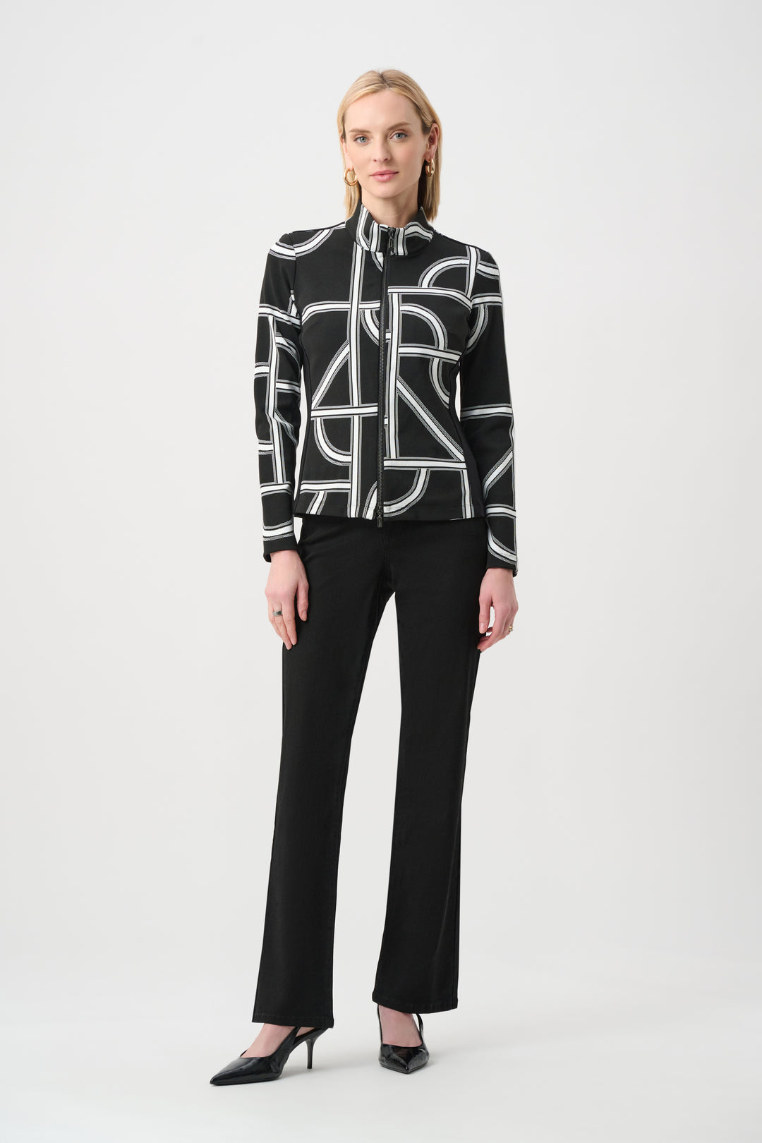 A woman standing in front of a light grey background wearing a black and white geometric print jacket and black pants. 