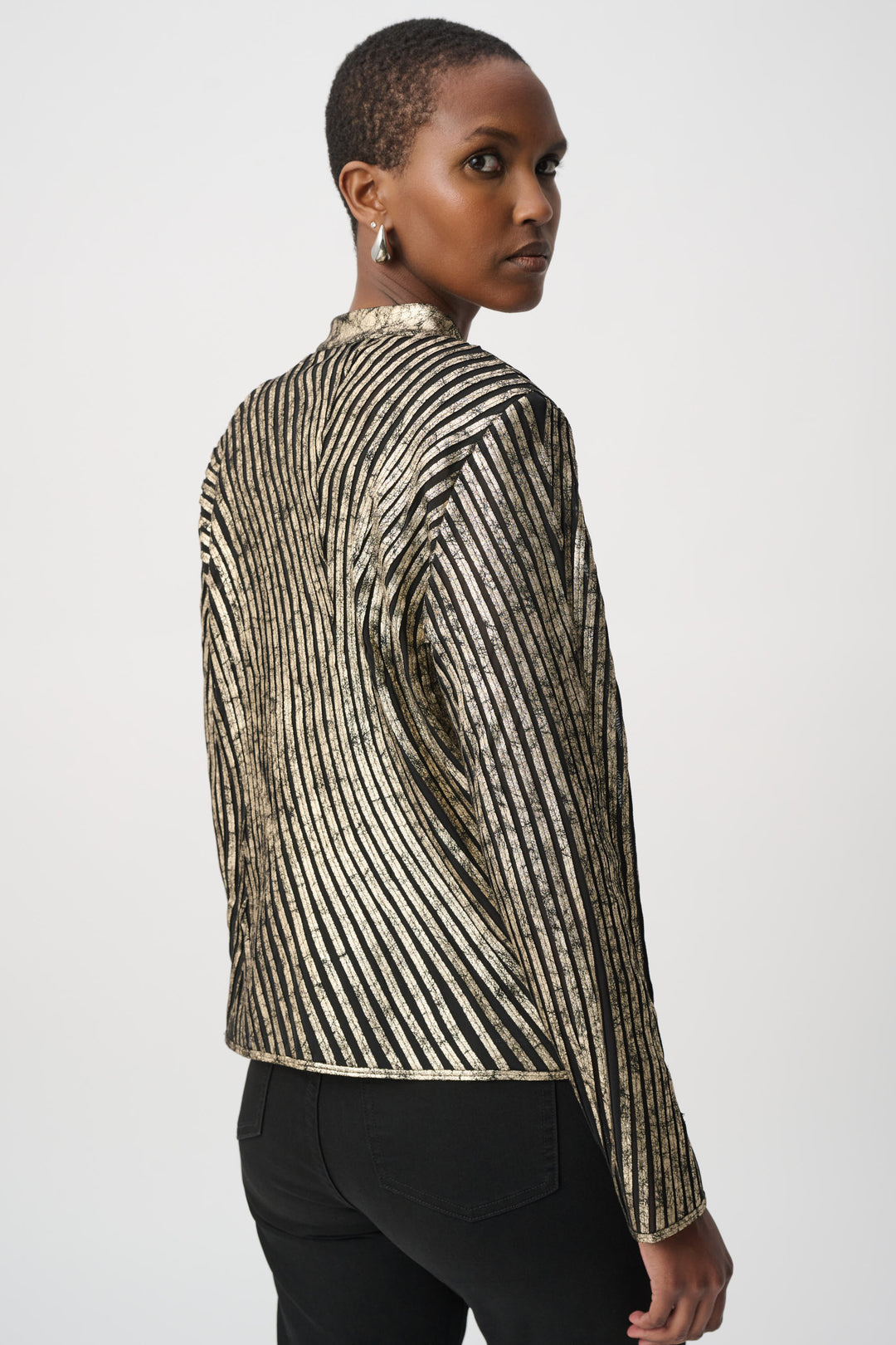 A woman shown from the back standing in front of a light grey background wearing a metallic gold jacket and black pants. 