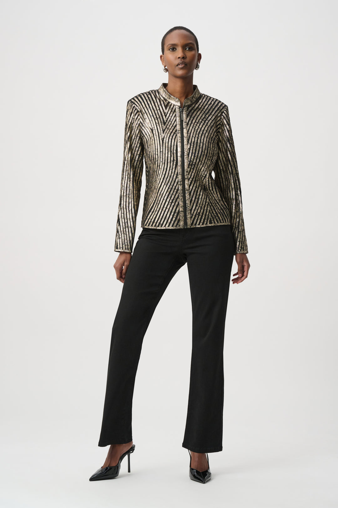 A woman standing in front of a light grey background wearing a metallic gold jacket and black pants. 