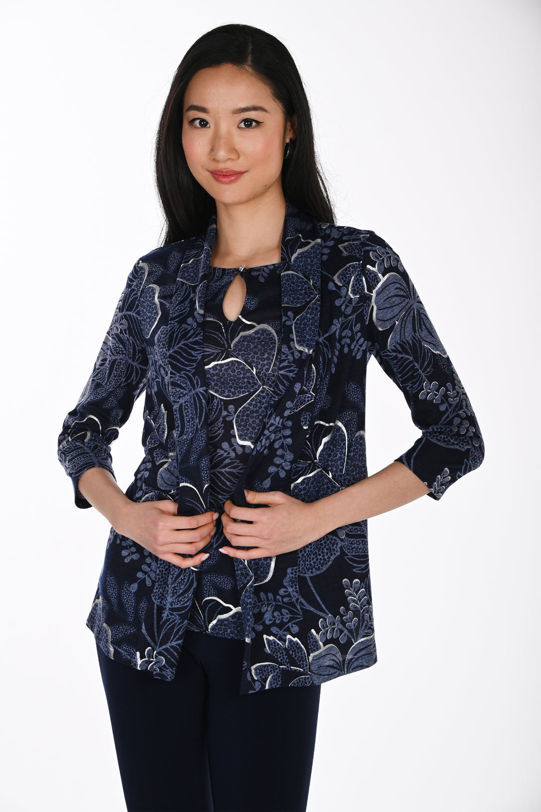 A woman wearing a navy, blue and silver floral patterned cardigan and navy pants standing in front of a light grey background. 