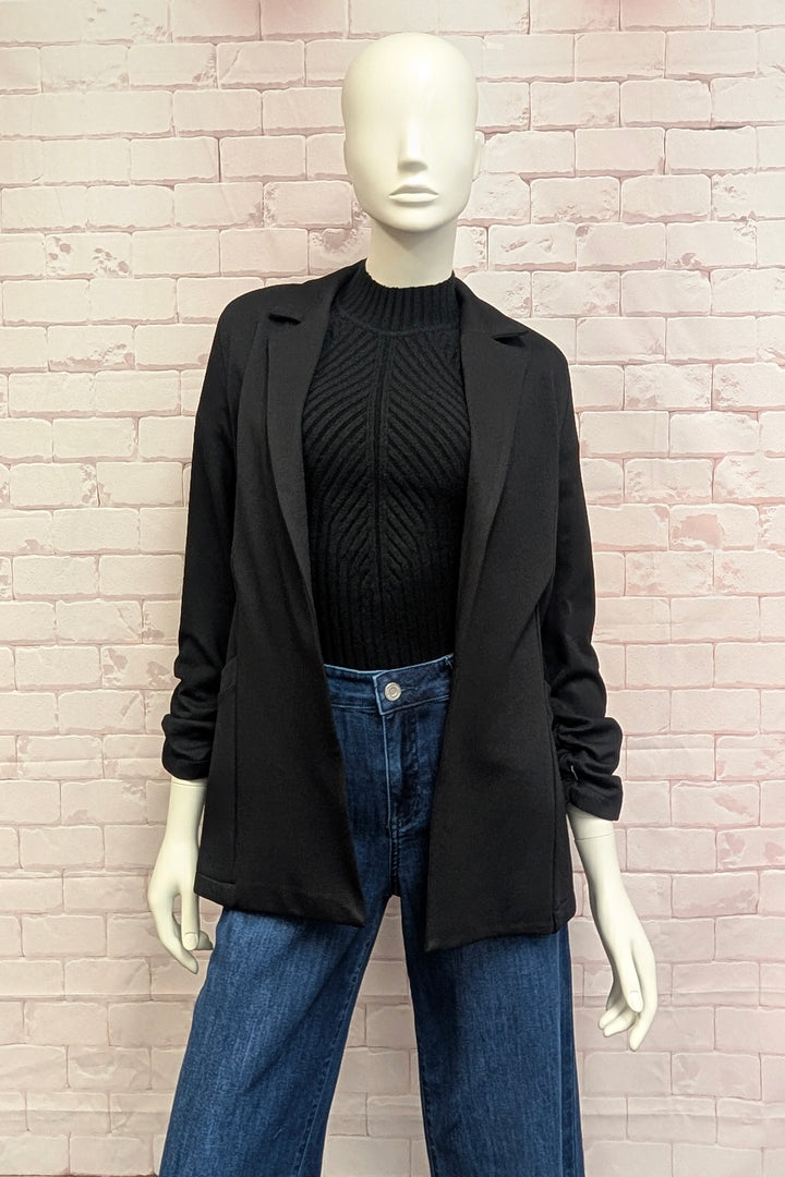 A mannequin standing in front of a white brick background modelling a black blazer with rouched sleeves. 
