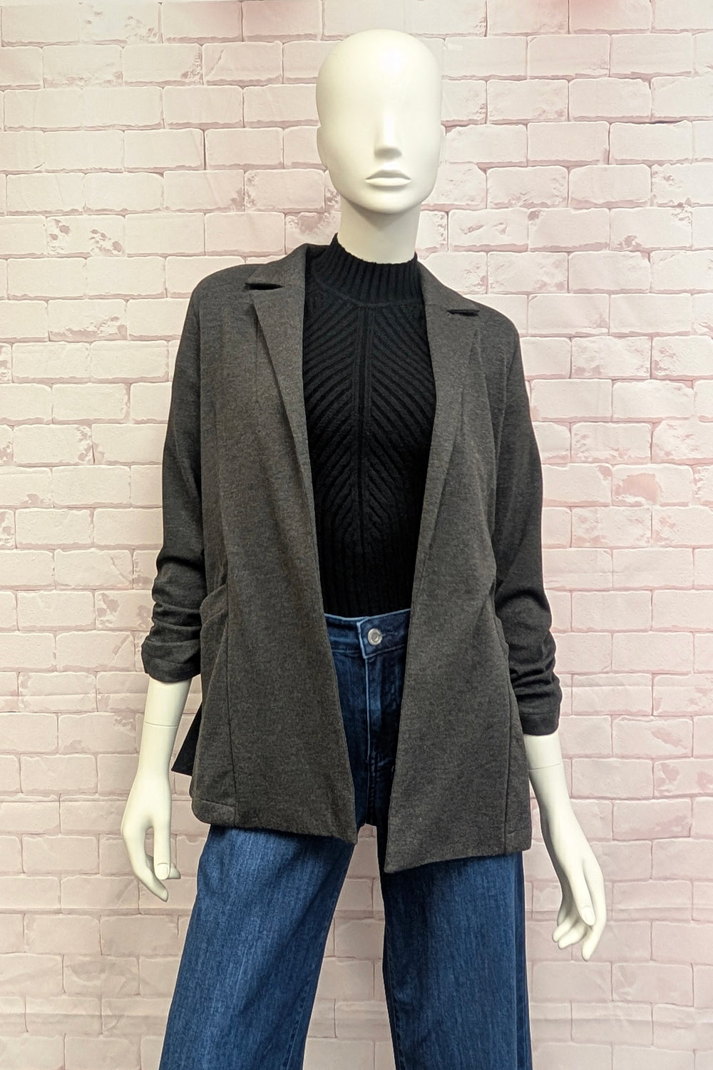 A mannequin standing in front of a white brick background modelling a grey blazer with rouched sleeves. 
