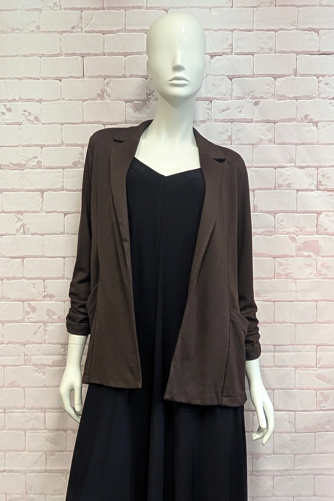 A mannequin standing in front of a white brick background modelling a brown blazer with rouched sleeves. 