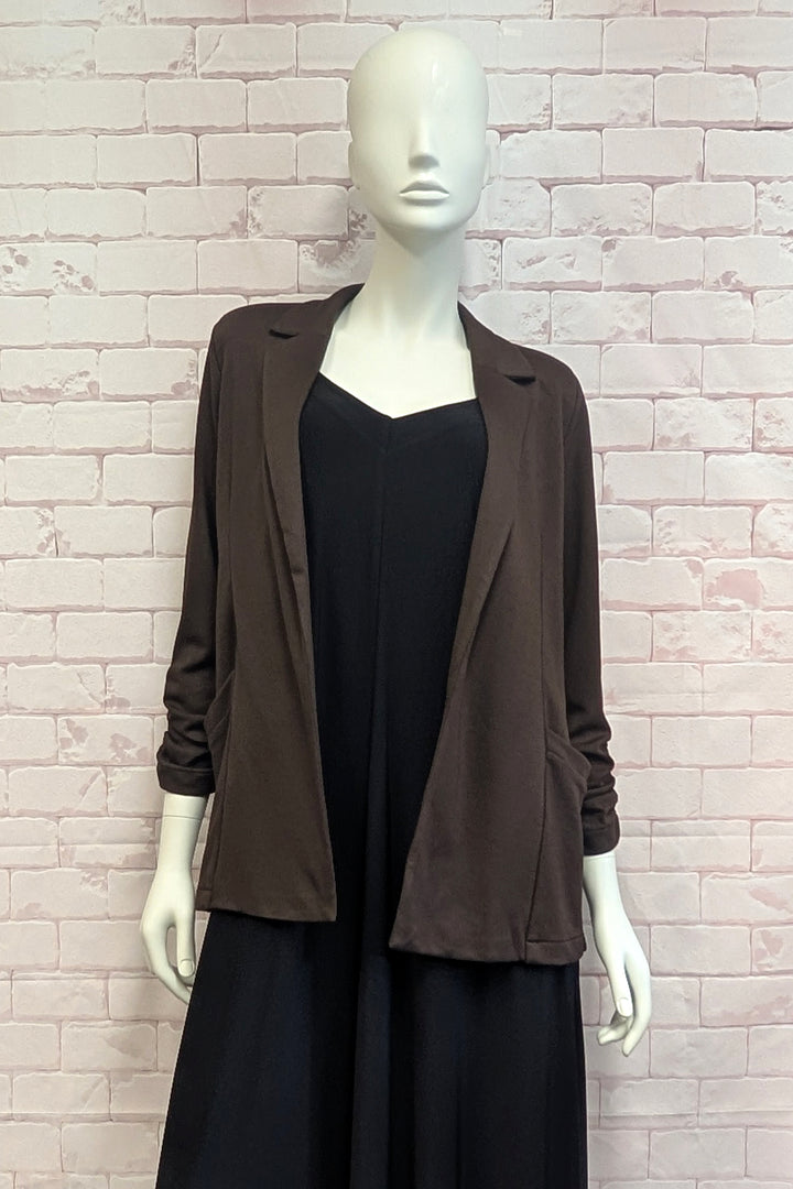 A mannequin standing in front of a white brick background modelling a brown blazer with rouched sleeves. 