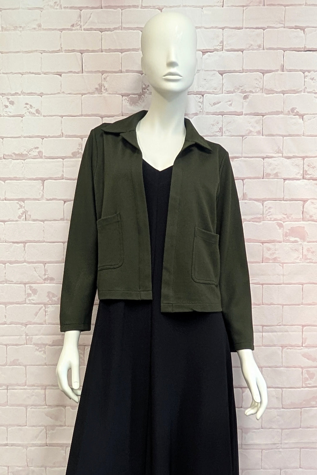 A mannequin standing in front of a white brick background modelling a cropped boxy olive green jacket. 
