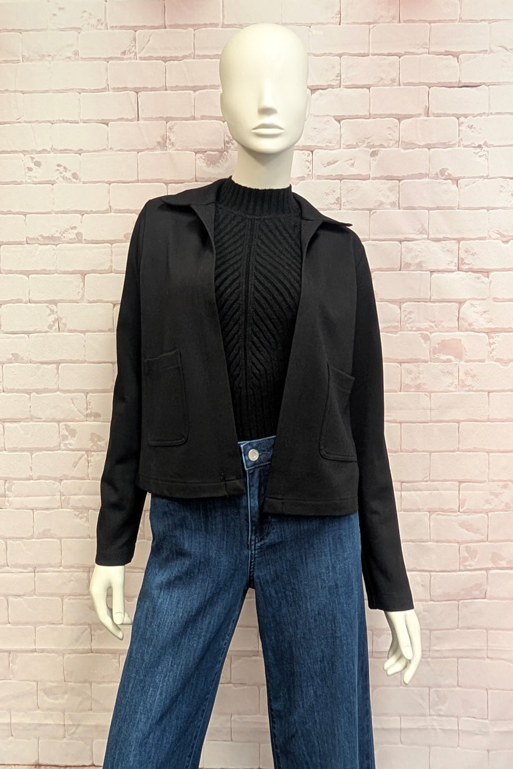 A mannequin standing in front of a white brick background modelling a cropped boxy black jacket. 
