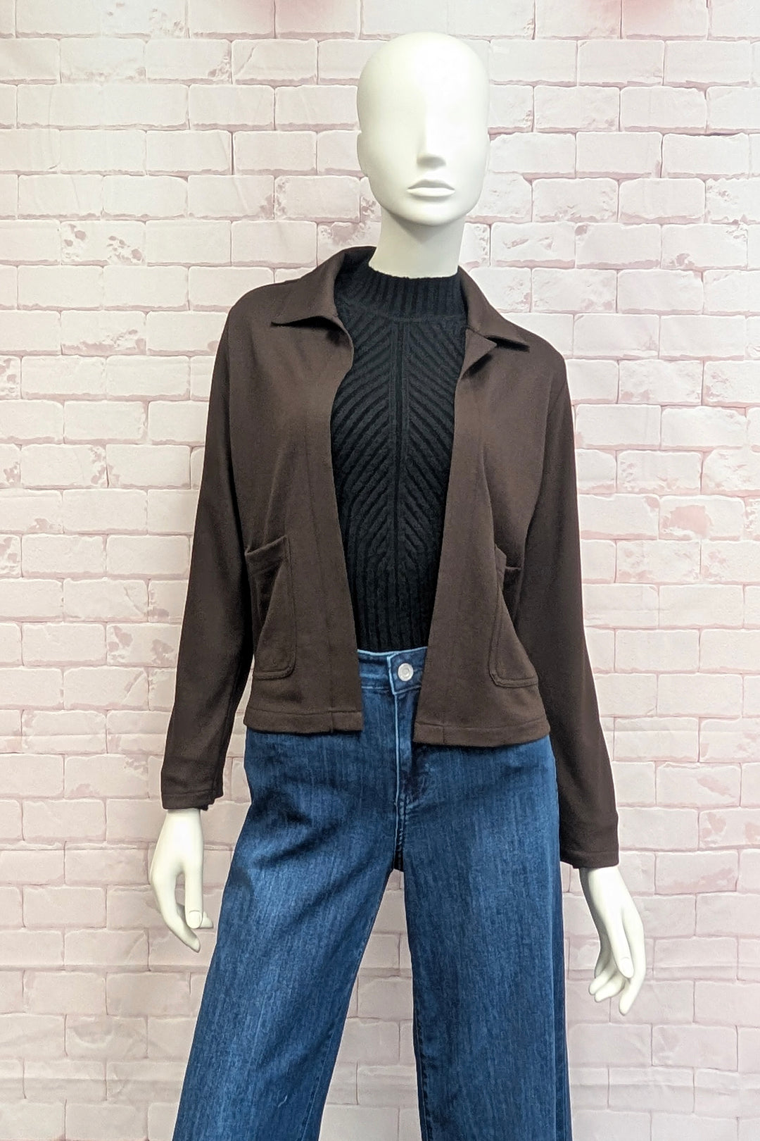 A mannequin standing in front of a white brick background modelling a cropped boxy brown jacket. 