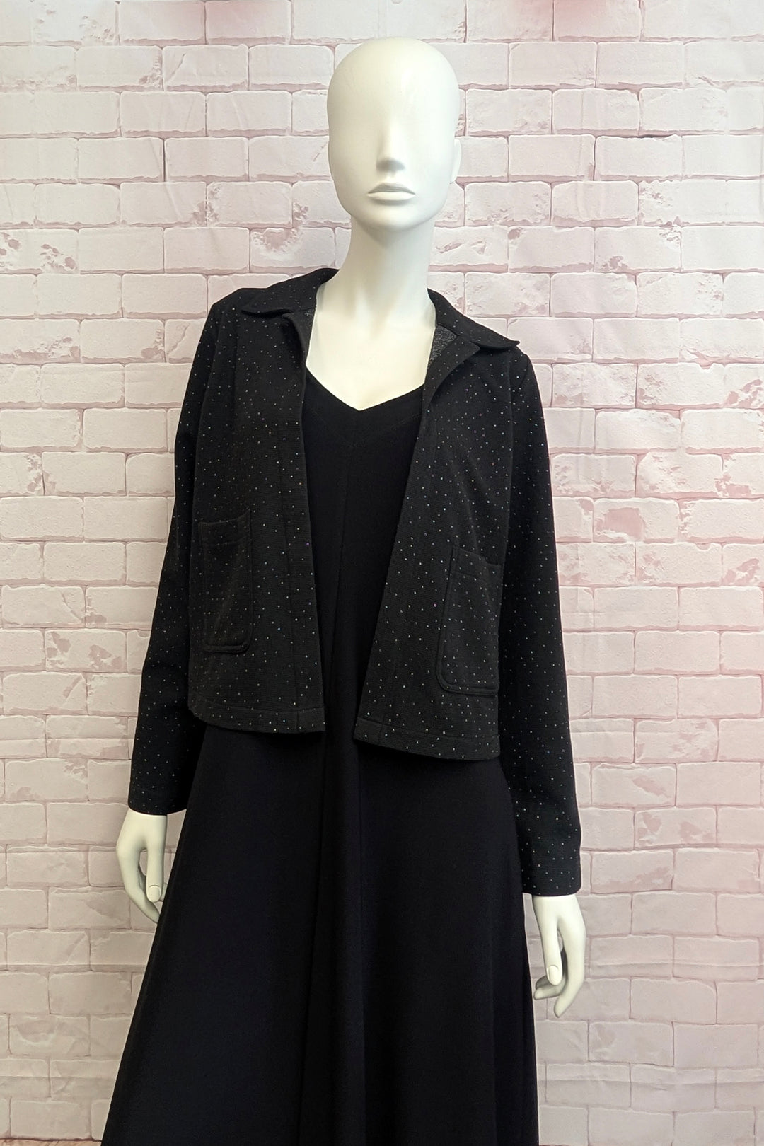 A mannequin standing in front of a light brick wall modelling a sparkly cropped jacket. 