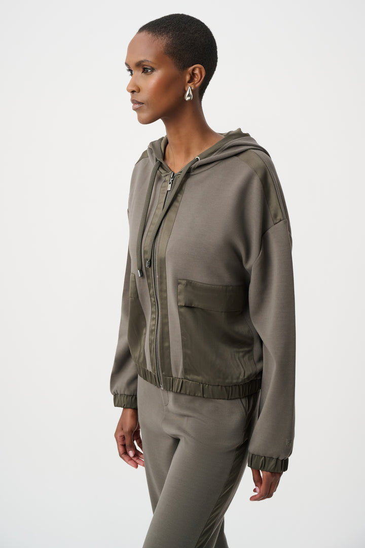 A woman standing in from of a light grey background wearing an olive coloured satin trim hoodie jacket and matching pants. 