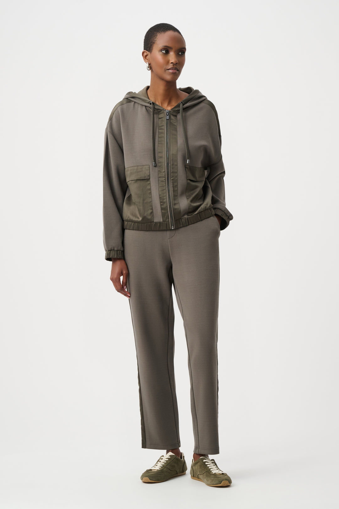 A woman standing in from of a light grey background wearing an olive coloured satin trim hoodie jacket and matching pants. 