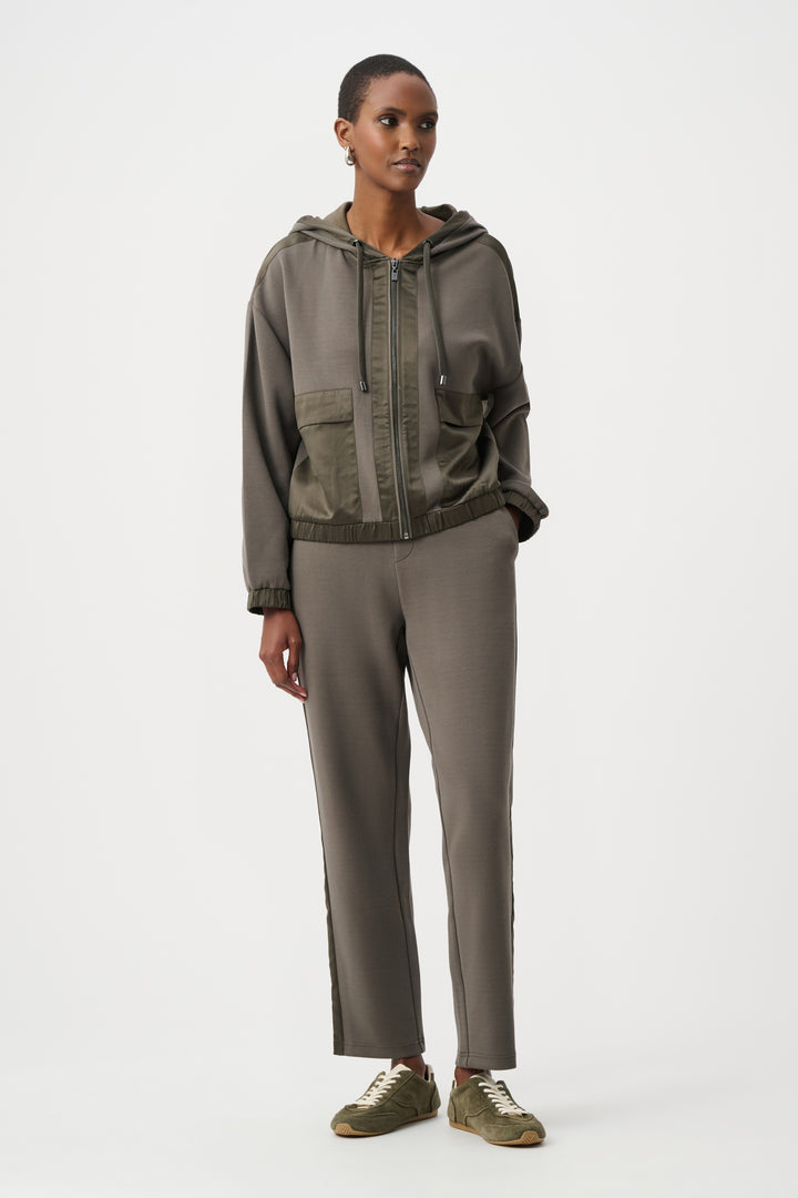 A woman standing in from of a light grey background wearing an olive coloured satin trim hoodie jacket and matching pants. 