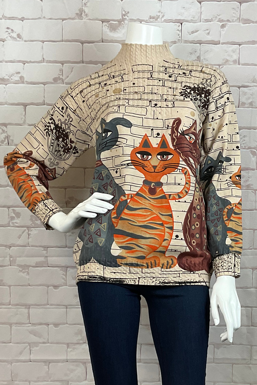 Ness Fall 2024 Made of light knit sweater fabric, this top features a ribbed contrast mock neck and full length sleeves. And the best part? Adorable cats on a biege 'bricks' background on the front and sleeves.