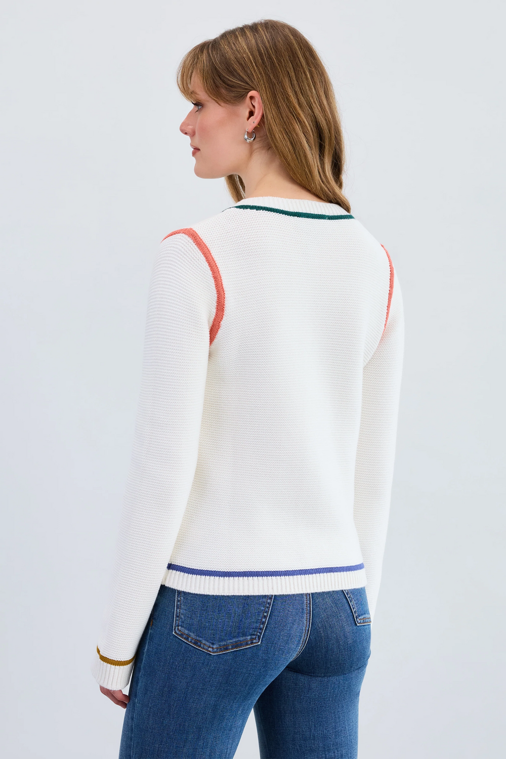 Woman wearing a white sweater with colorful accents and blue jeans on a light gray background