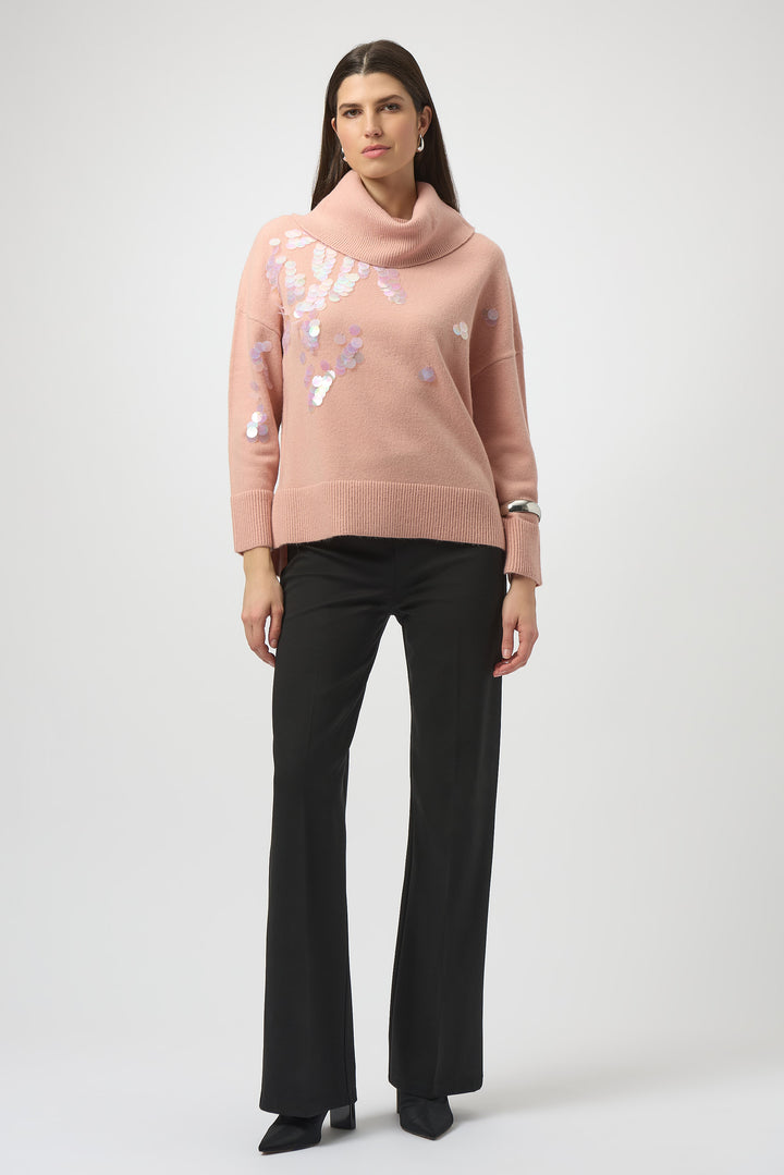 Woman wearing a pink sweater with floral embellishments and black pants on a white background