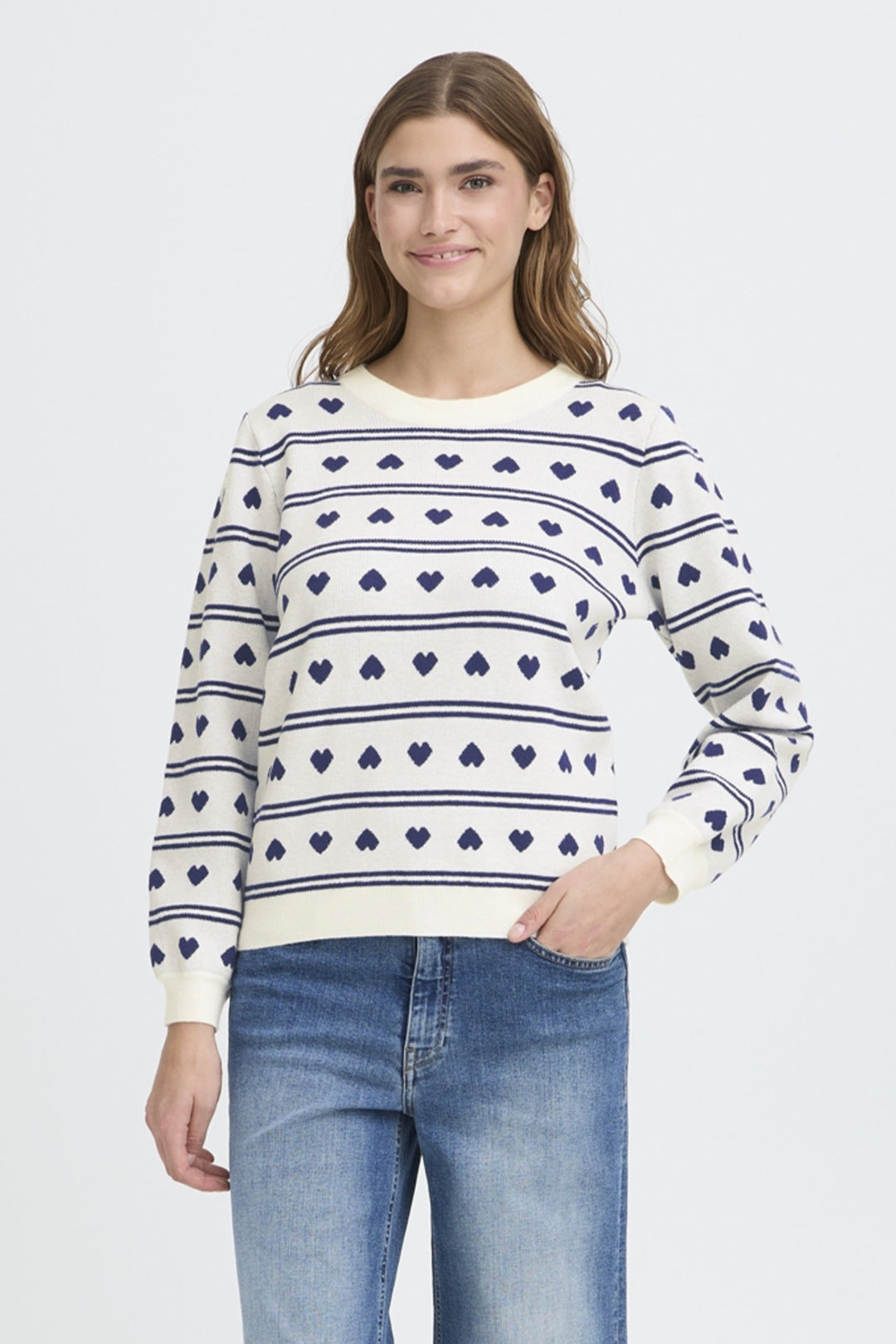 Woman wearing a white sweater with blue heart patterns and jeans on a light gray background