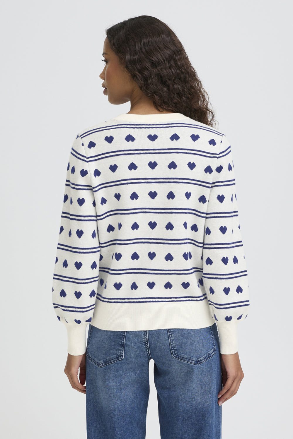 Woman wearing a white sweater with blue heart patterns and blue jeans on a light gray background